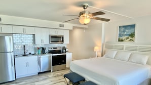 Een kleine hotelkamer met kitchenette, een bed met wit beddengoed, een plafondventilator en een schilderij aan de muur.