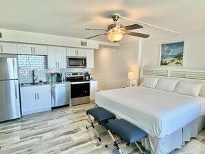 A compact hotel room with a kitchenette, a bed with white bedding, a ceiling fan, and a painting on the wall.