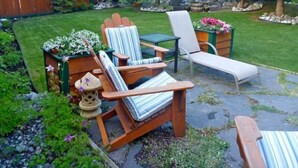 A backyard with a wooden deck, two Adirondack chairs, a small table, and a garden area with plants and a wooden fence. - Private studio appartment with great outdoor area (Canmore)