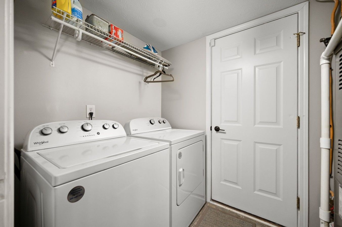 Une buanderie équipée de deux machines à laver et de deux sèche-linge Whirlpool, d’une étagère en métal contenant des produits de nettoyage et d’une porte blanche.