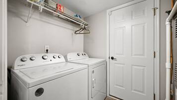 Une buanderie équipée de deux machines à laver et de deux sèche-linge Whirlpool, d’une étagère en métal contenant des produits de nettoyage et d’une porte blanche.