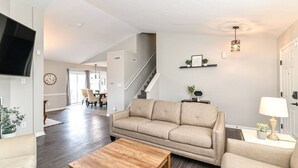 A living room with a sofa, coffee table, and a TV mounted on the wall. There is a staircase leading to another level, and a dining area in the background.