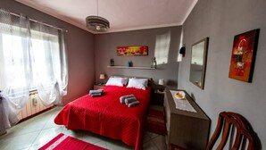 Quarto com colcha vermelha, mesa de cabeceira com abajur, cadeira e prateleira na parede com objetos decorativos.