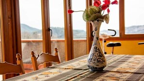 Mesa de jantar com toalha florida, vaso com flores vermelhas e vista para montanhas através de grandes janelas.