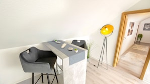 Une pièce moderne et minimaliste, avec un coin repas comprenant deux chaises grises et une table en marbre. Un lampadaire à abat-jour jaune est placé près de l'entrée.