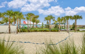 A sandy beach area with palm trees, a volleyball net, and a building with a red chair in the background.