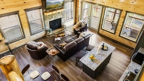 A cozy cabin interior with a stone fireplace, wooden walls, and large windows. - Luxury Indoor Pool Cabin Near Gatlinburg - Hot Tub, Game Room & Fire Pit (Sevierville)