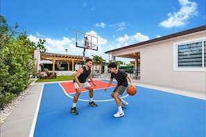 Een buitenbasketbalveld met twee spelers, een overdekt zitgedeelte en een gebouw met ramen.