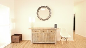 Un mueble de madera con cajones, un espejo redondo y una silla blanca en una habitación con suelo de madera y paredes blancas.