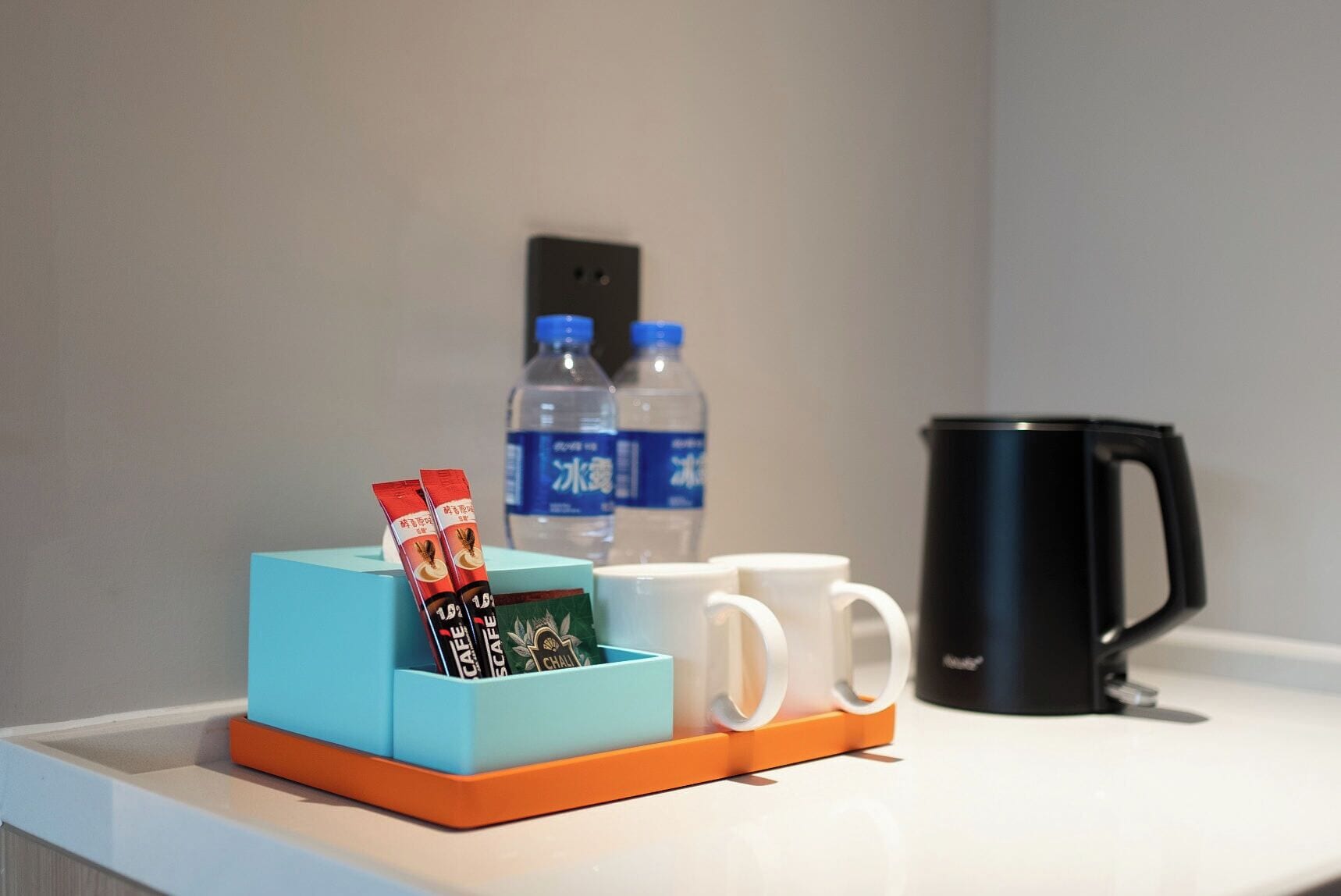 Habitación de hotel con una pequeña mesa que sostiene una caja azul con una cafetera, dos botellas de agua y una taza negra.