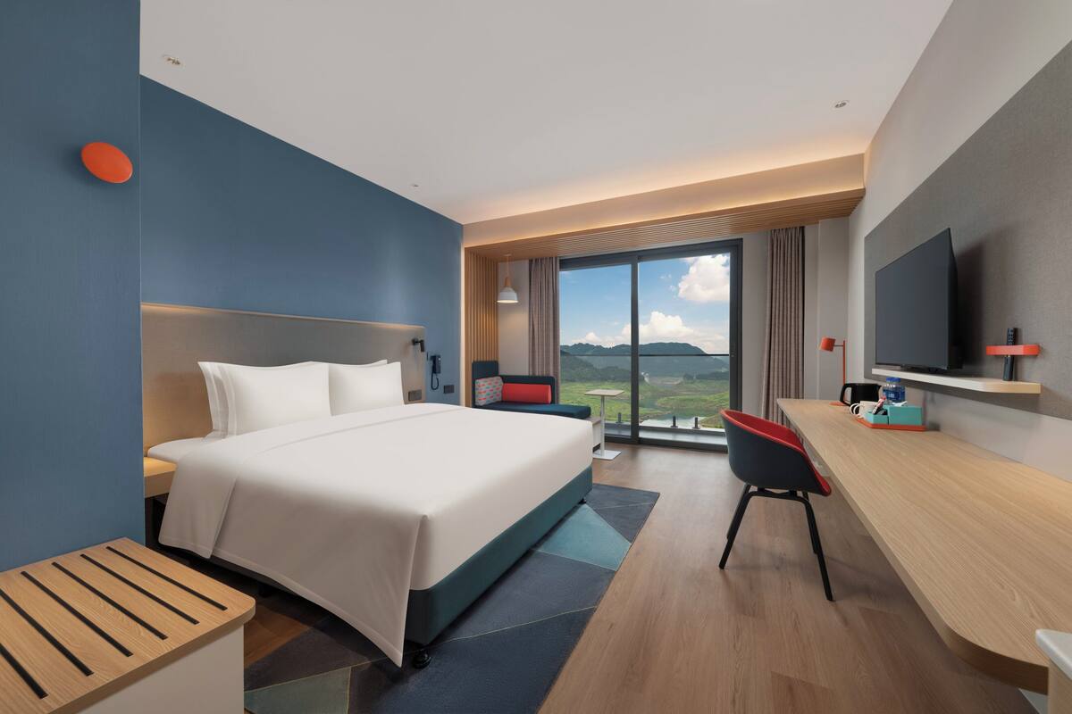 A modern hotel room with a large bed, a desk, a chair, a TV, and a view of the mountains.
