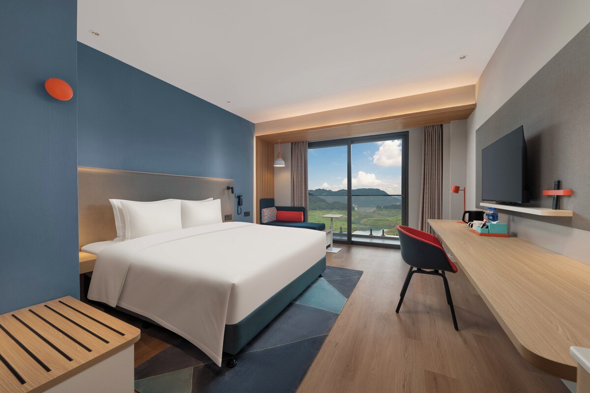 A modern hotel room with a large bed, a desk, a chair, a television, and a view of the outdoors.