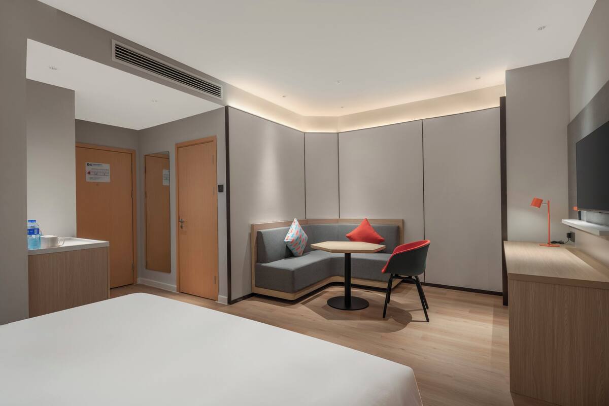 A modern hotel room with a bed, a small round table with two chairs, a sofa with cushions, a TV, and a desk with a chair.
