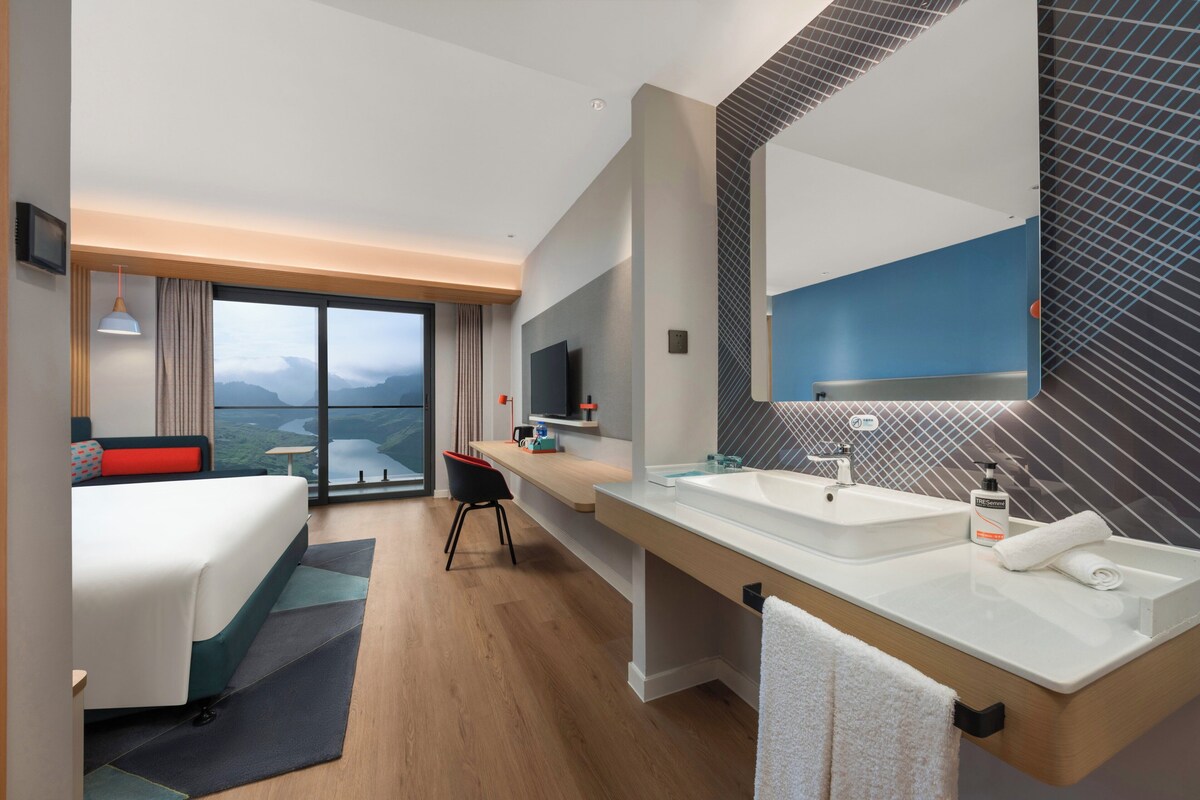 A hotel room with a large bed, a desk, a chair, a TV, a bathroom with a sink and mirror, and a view of mountains through the window.