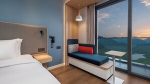A modern hotel room with a bed, a small table, a sofa with cushions, a phone, and a large window with a view of the mountains.