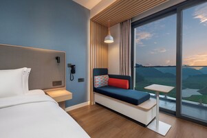 Una habitación de hotel moderna con cama, una mesita, un sofá con cojines, un teléfono y un ventanal con vistas a las montañas.