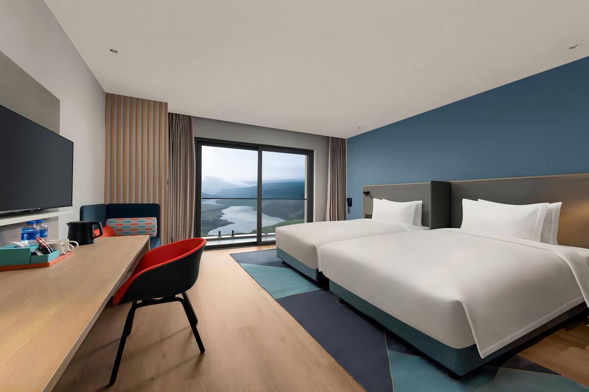 A hotel room with a large bed, a desk with a chair, a flat-screen TV, and a view of a mountainous landscape through the window.