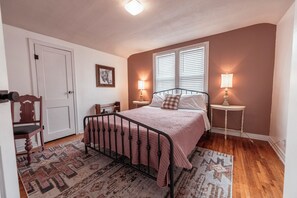 Une chambre à coucher avec un lit, deux tables de chevet avec des lampes, une chaise, une porte et une fenêtre avec des stores.
