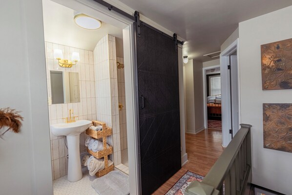 Une salle de bain moderne avec un lavabo sur colonne blanc, un grand miroir et une porte coulissante noire donnant sur une chambre.