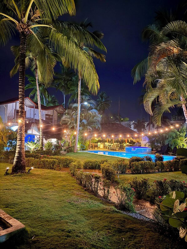 A nighttime view of a resort with a swimming pool, palm trees, and string lights.