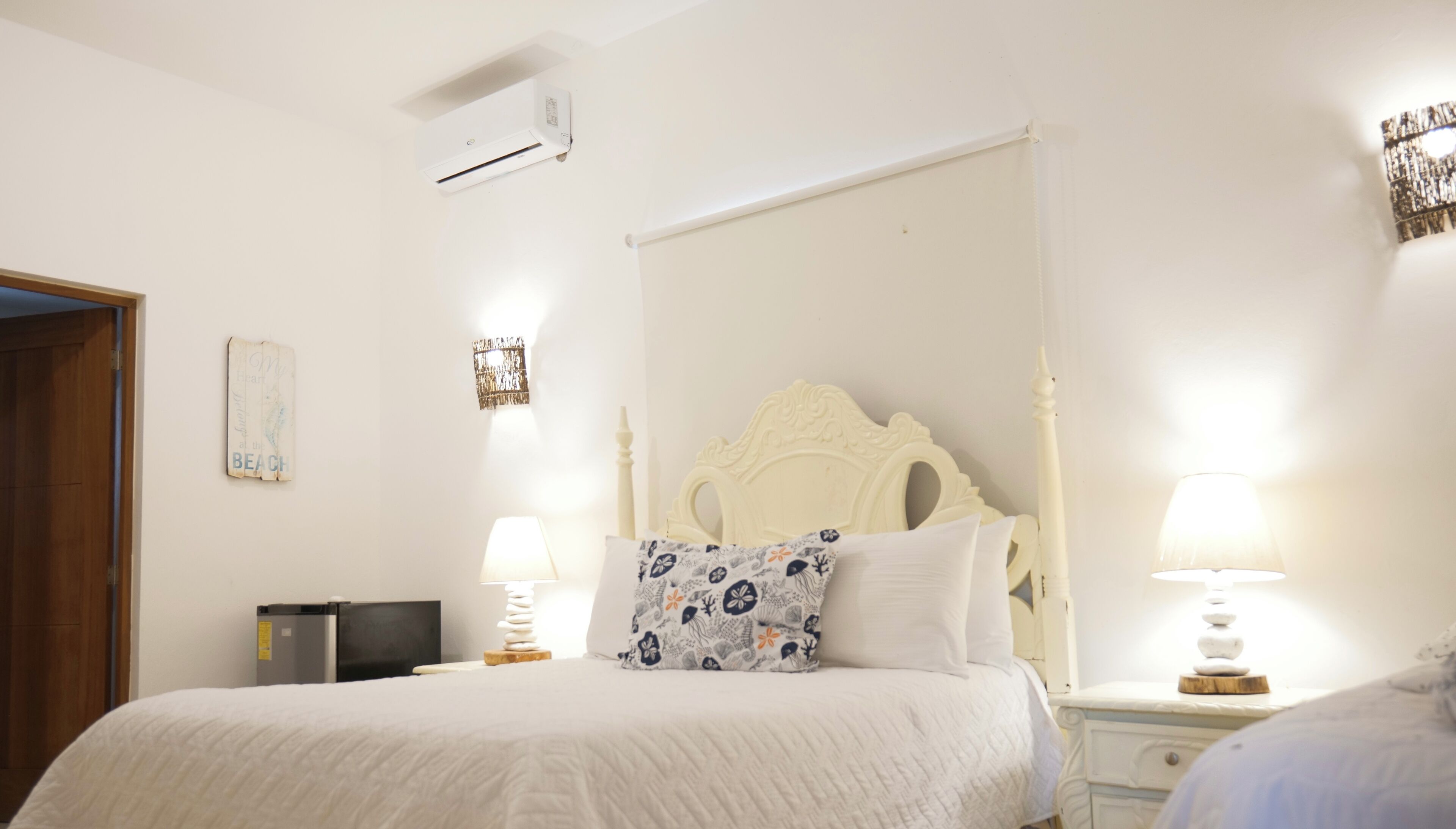 A bedroom with a bed, two bedside tables with lamps, a nightstand, a wall-mounted air conditioner, and a decorative mirror.