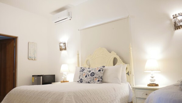 Une chambre à coucher comprenant un lit, deux tables de chevet avec des lampes, une commode, un climatiseur fixé au mur et un miroir décoratif.