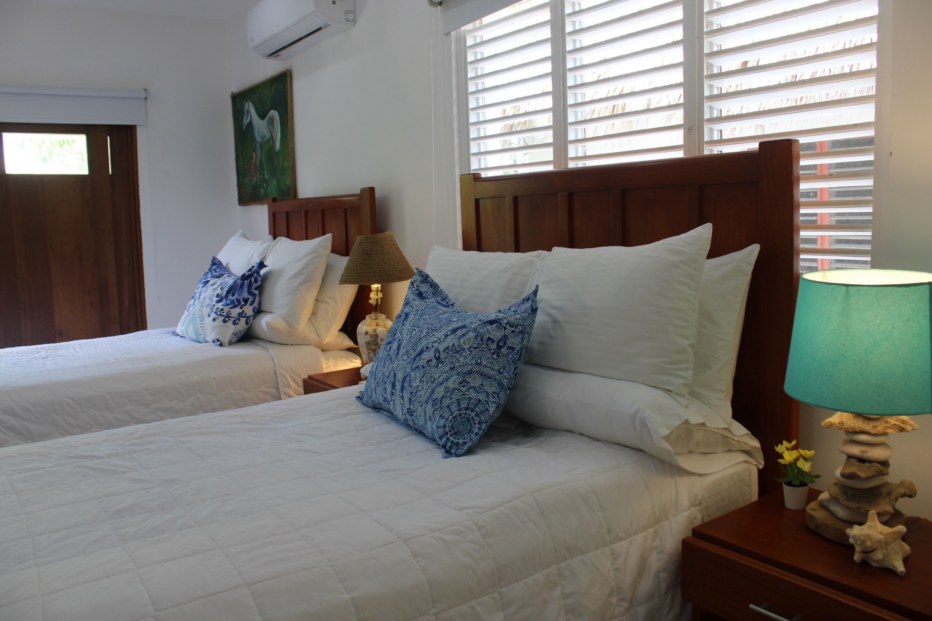 A hotel room with two beds, each with white bedding and blue patterned pillows. There is a wooden headboard, a nightstand with a lamp, and a window with blinds.