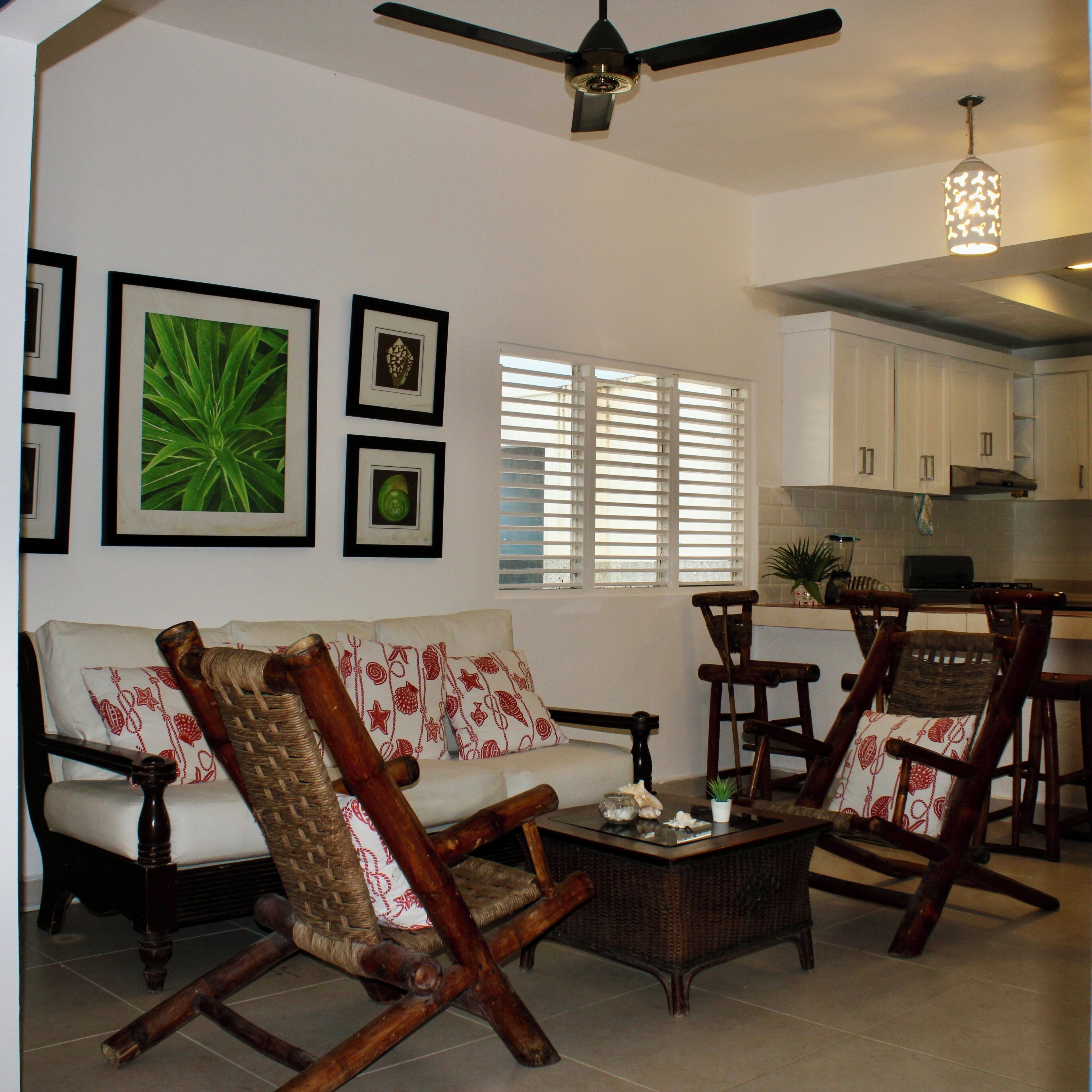 A living room with a sofa, two wooden chairs, a coffee table, and a ceiling fan. There are framed pictures on the wall and a window with blinds.