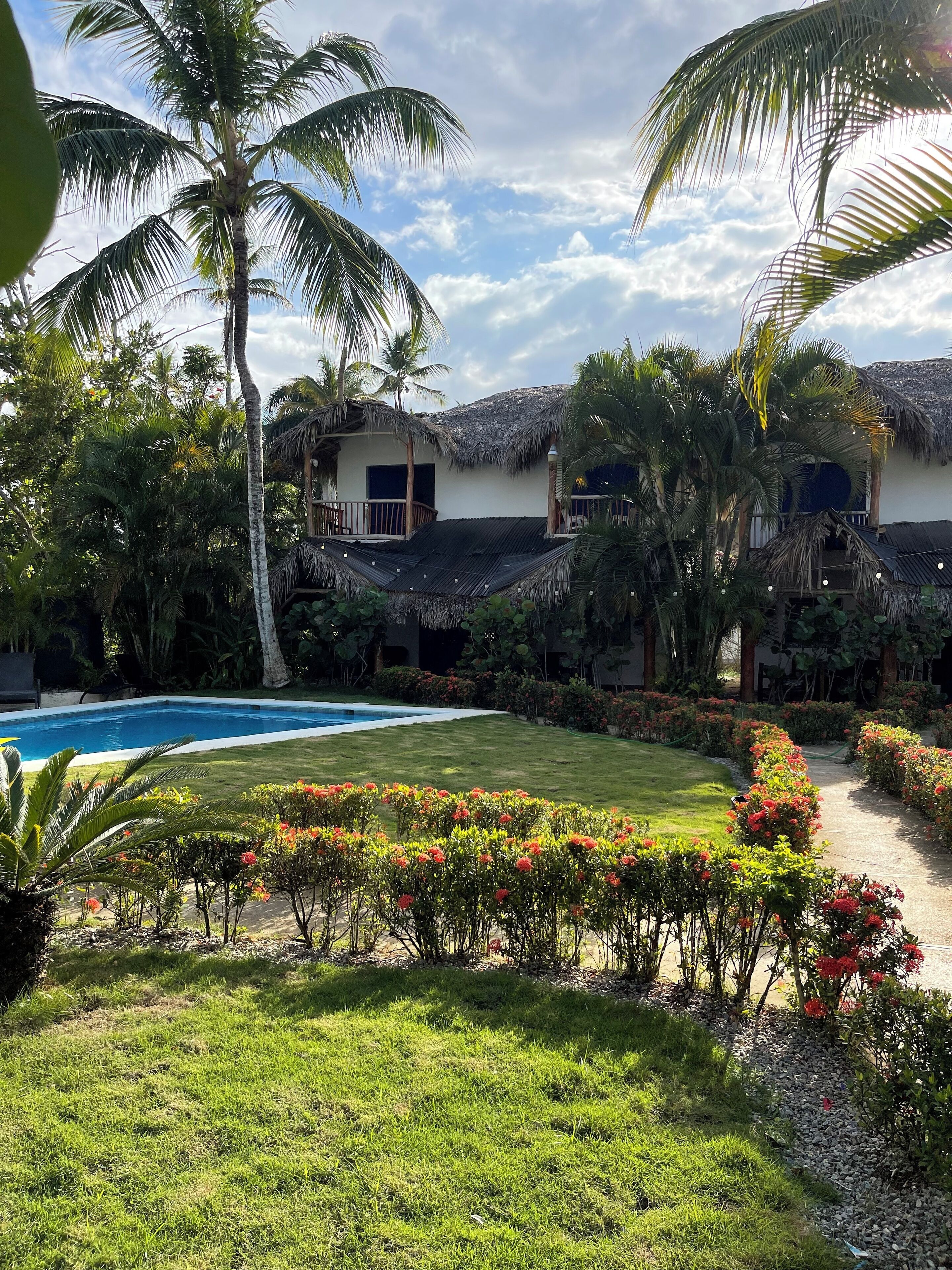 A pool surrounded by lush greenery and flowering plants, with a thatched-roof building in the background.