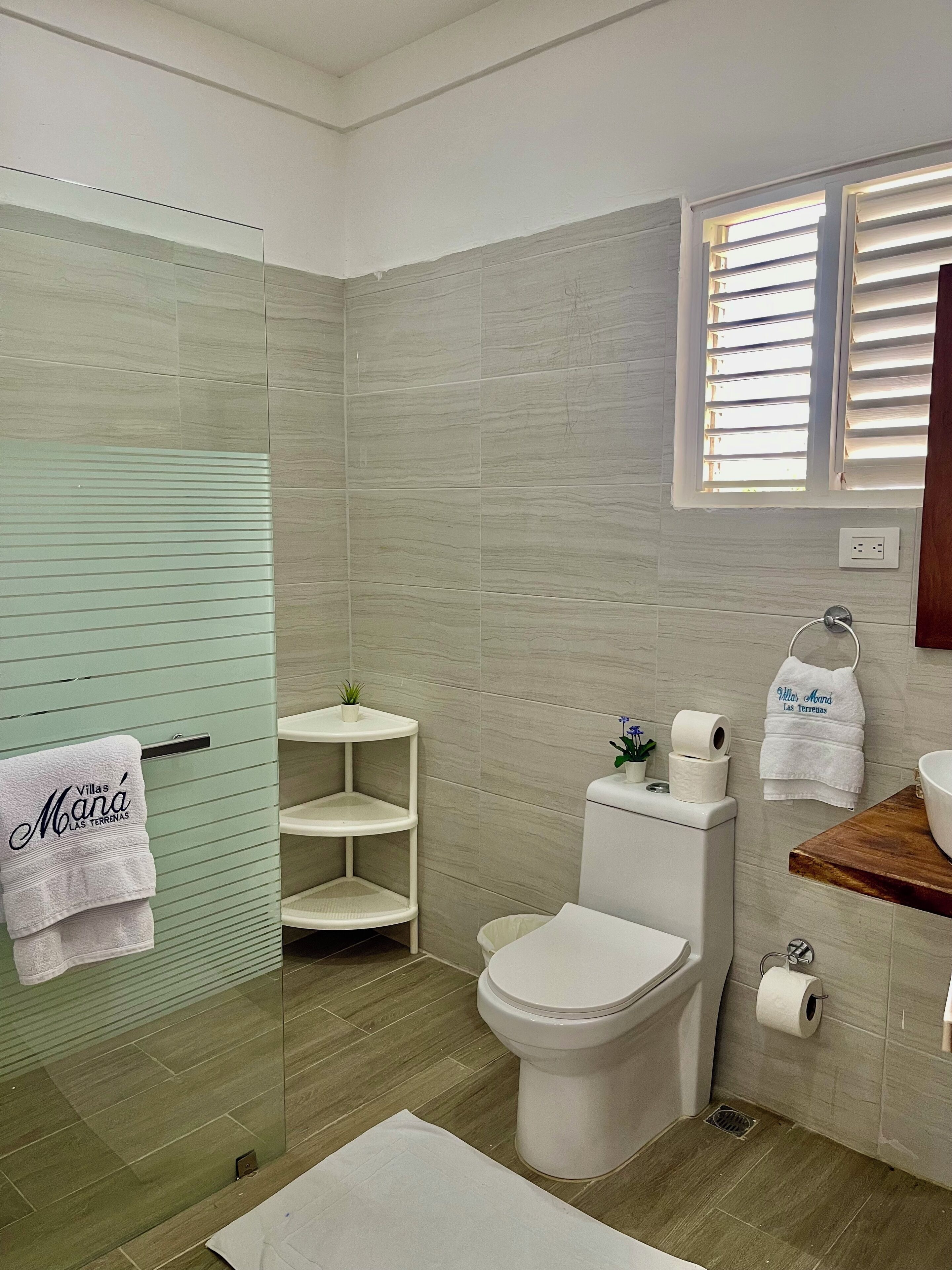 Un baño moderno con cabina de ducha de vidrio, inodoro y tocador de madera, y ventana con persianas.