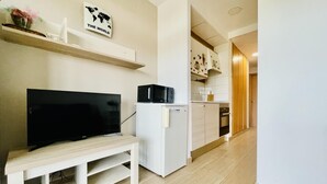 A modern living room with a flat-screen TV, a small white fridge, and a wooden bench. A world map sign is on the wall, and a kitchen is visible in the background.