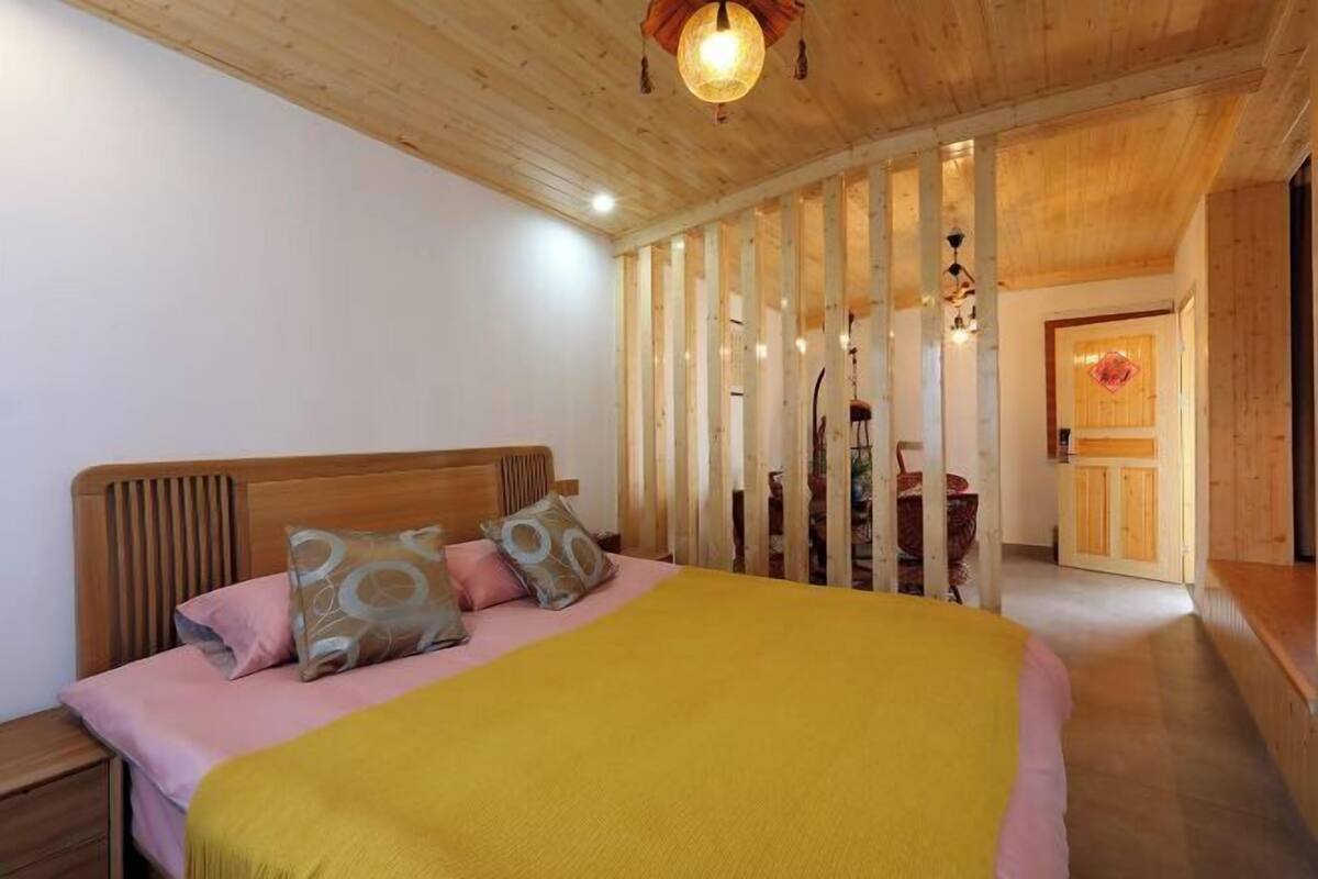 Une chambre à coucher avec une tête de lit en bois, un grand lit recouvert d’une couverture jaune et une cloison en bois avec des lattes verticales.