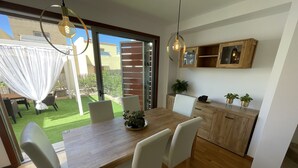En matplats med ett träbord och vita stolar, ett litet köksområde med träskåp och en utsikt över en grön gräsmatta och en byggnad genom en glasdörr.