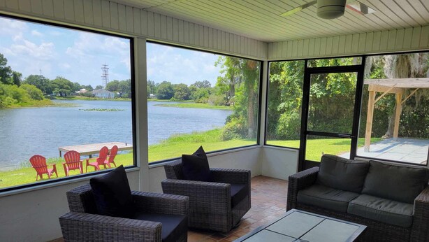 A sunroom with wicker furniture, a glass table, and a view of a lake and greenery. - Charming Lake Home (Lake City)