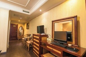 Habitación de hotel con un escritorio de madera, una computadora y un espejo. Hay una silla y una mesita con un florero. El piso es de madera y la pared tiene un diseño.