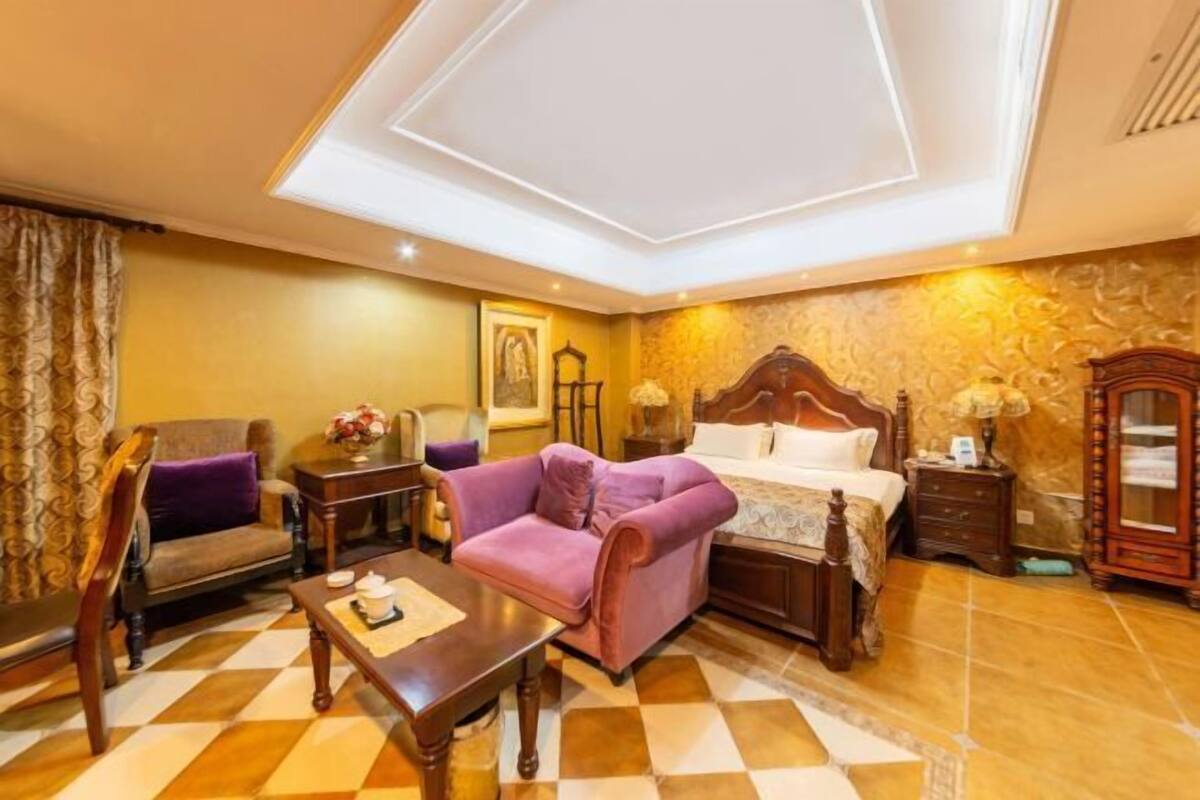 Una habitación de hotel con piso a cuadros, una cama con cabecera de madera, un sillón morado, una mesita de noche de madera y una lámpara.