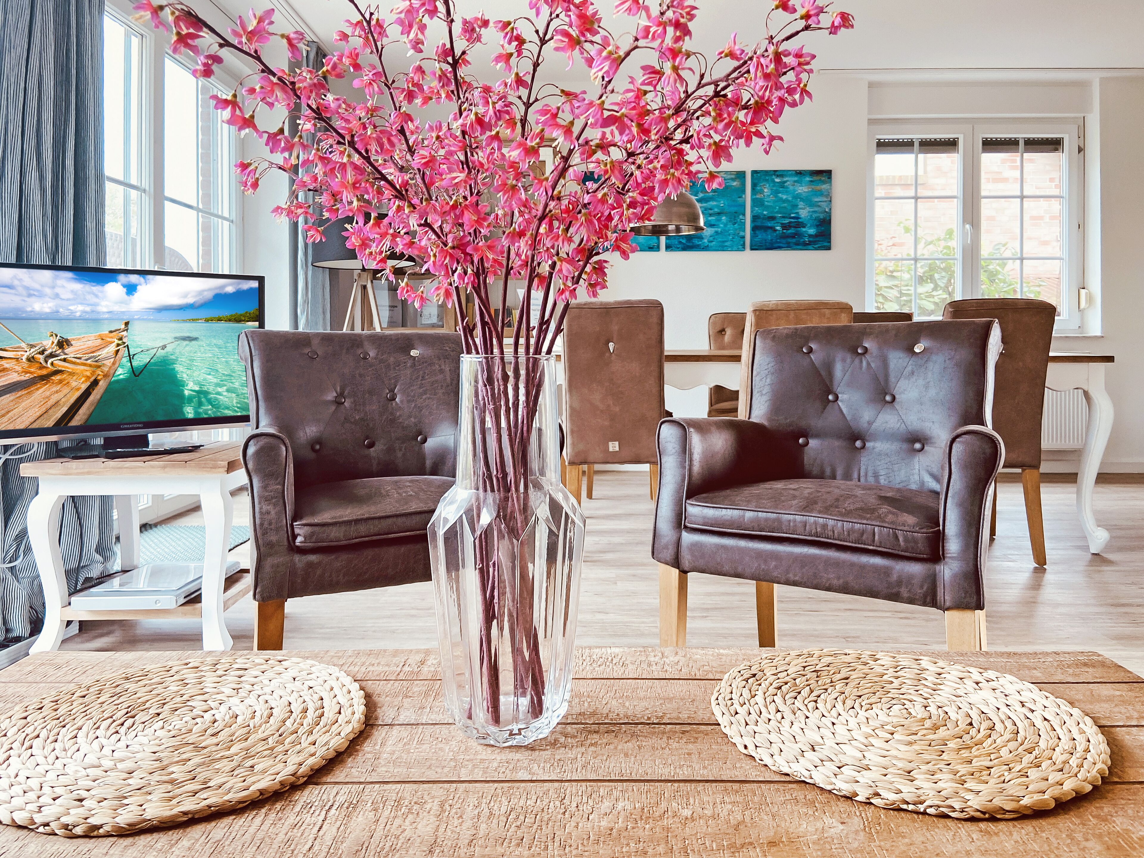 Ein Wohnzimmer mit zwei braunen Ledersesseln, einer klaren Vase mit rosa Blumen, einem Fernseher und einem Essbereich im Hintergrund.