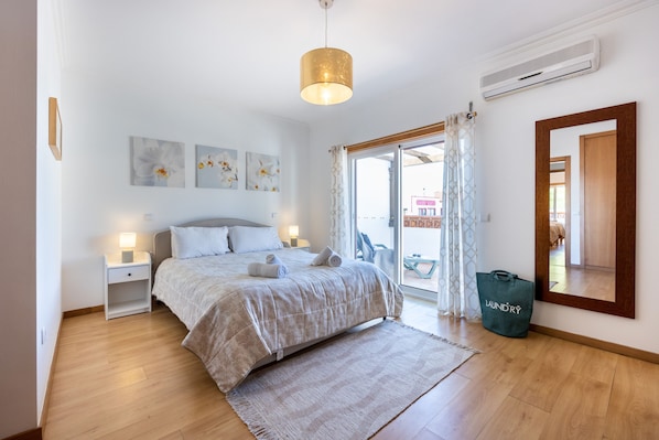 A bedroom with a bed, bedside tables, a hanging light fixture, a large window with curtains, and a mirror.