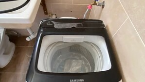 Une machine à laver Samsung dans une salle de bain équipée de toilettes et où l’on peut voir une tuyauterie.
