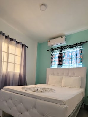Une chambre à coucher avec un lit doté d’une tête de lit capitonnée, une fenêtre ornée de rideaux à motifs et un appareil de climatisation placé au-dessus de la fenêtre.