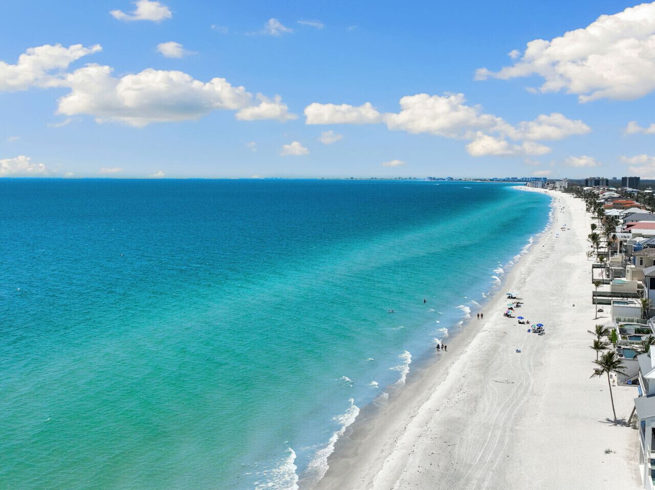 Une plage à l’eau turquoise et limpide, un rivage sablonneux et des bâtiments qui bordent la côte.