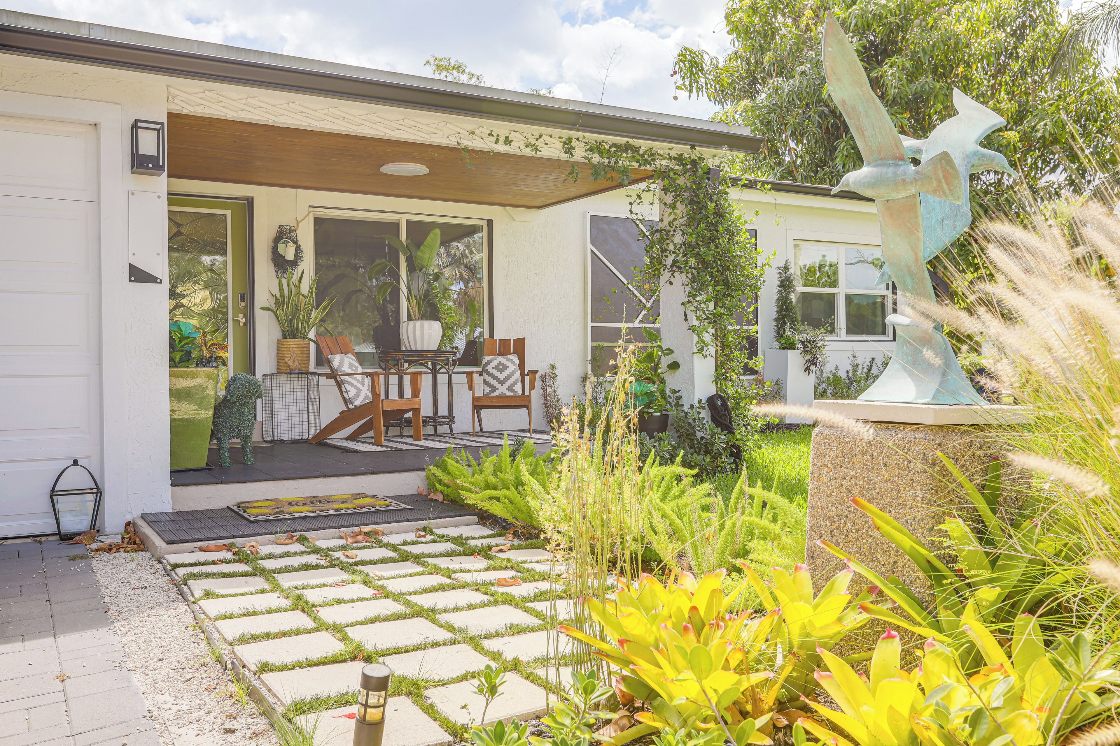 A house with a covered porch, a garden with various plants, and a sculpture.