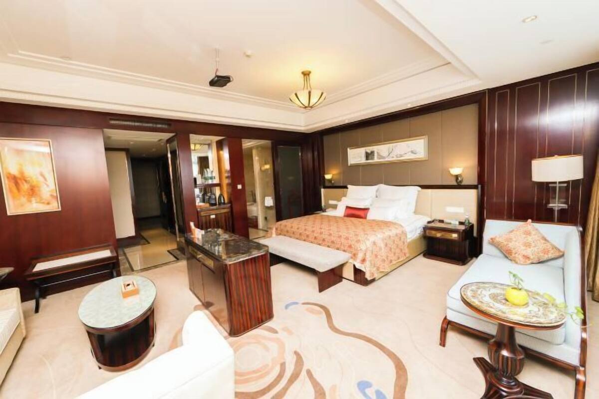 Habitación de hotel con una cama grande, una zona de estar con sofá y sillón, una mesa redonda pequeña y un banco.