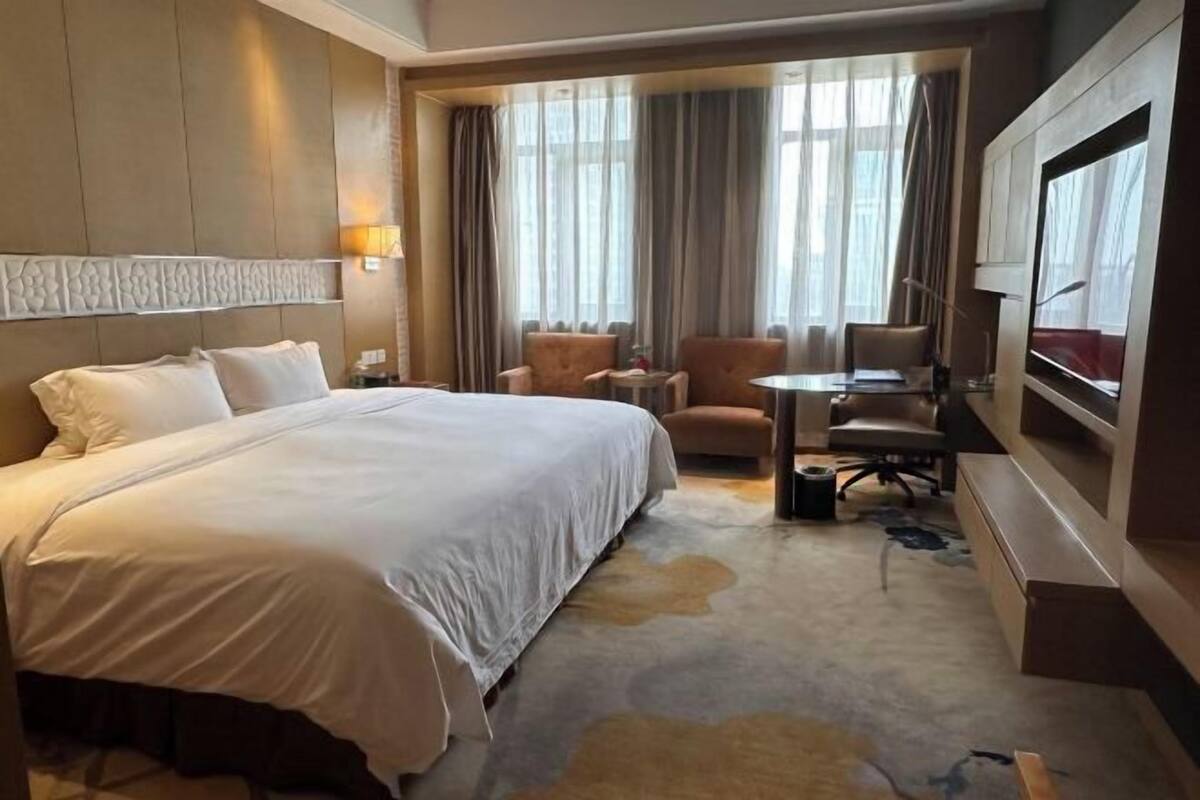 Habitación de hotel con una cama grande, un escritorio, dos sillas, un televisor y vistas a la ciudad a través de amplias ventanas.