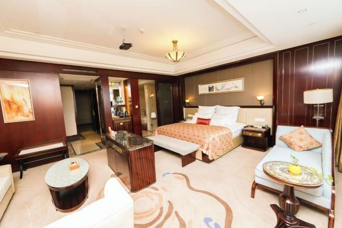 A hotel room with a bed, a sofa, a small table, a bench, a mirror, and a chandelier.