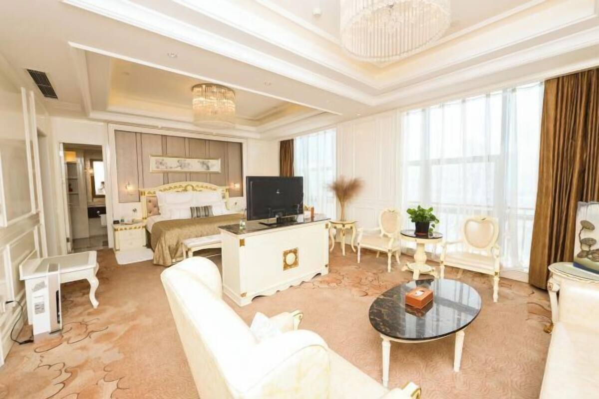 Amplia habitación de hotel con una cama grande, televisión, zona de comedor y sala de estar con candelabro.