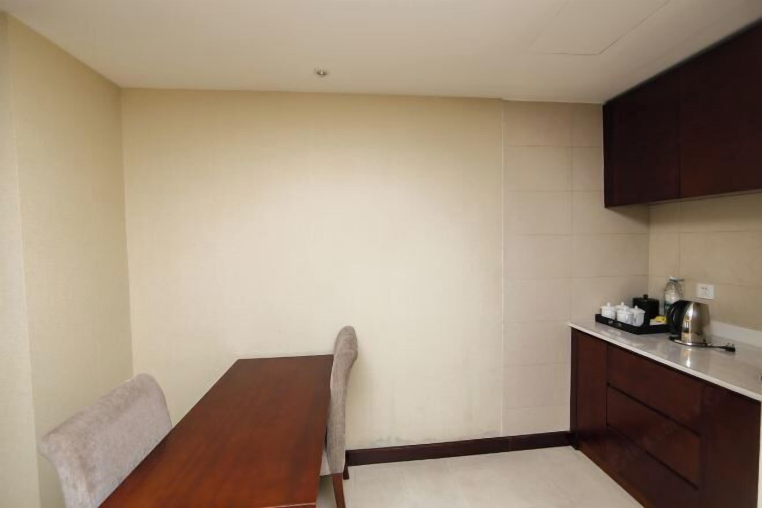 Una habitación pequeña y limpia con una mesa de madera, una silla, una kitchenette con mostrador y armarios, y una pared sencilla.