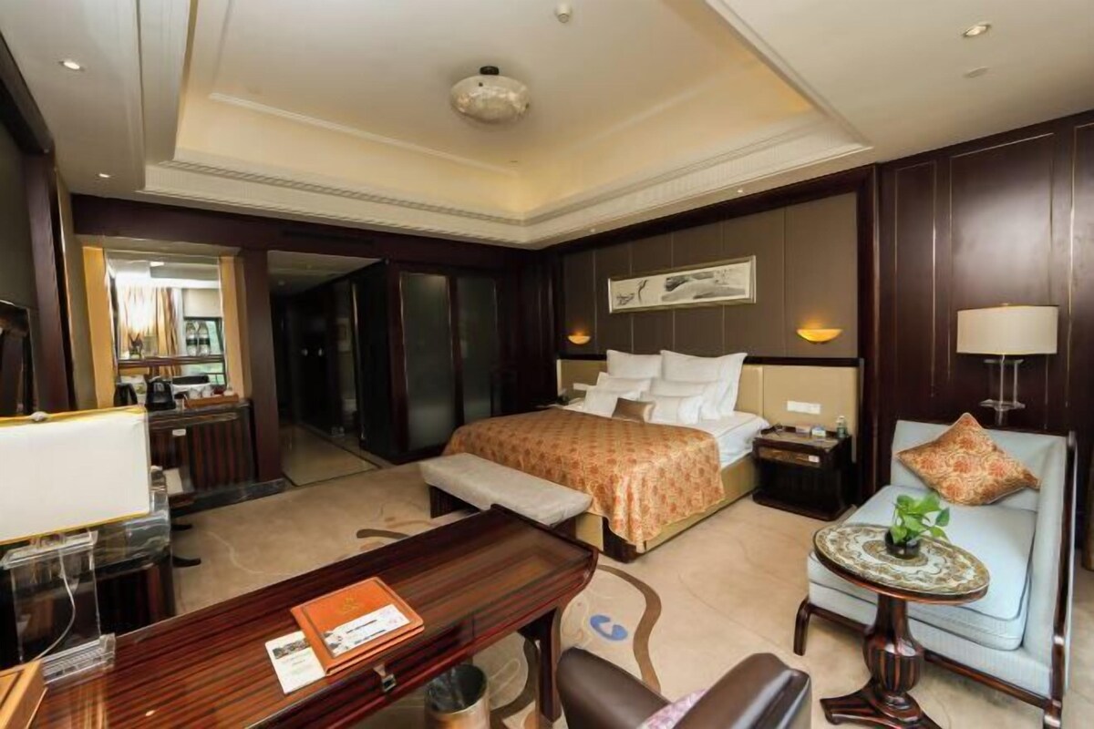 A hotel room with a bed, a desk, a chair, a sofa, a lamp, and a small table with a plant.