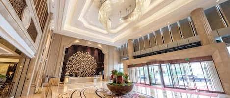 A spacious lobby with a central reception desk, a chandelier, and a large window.