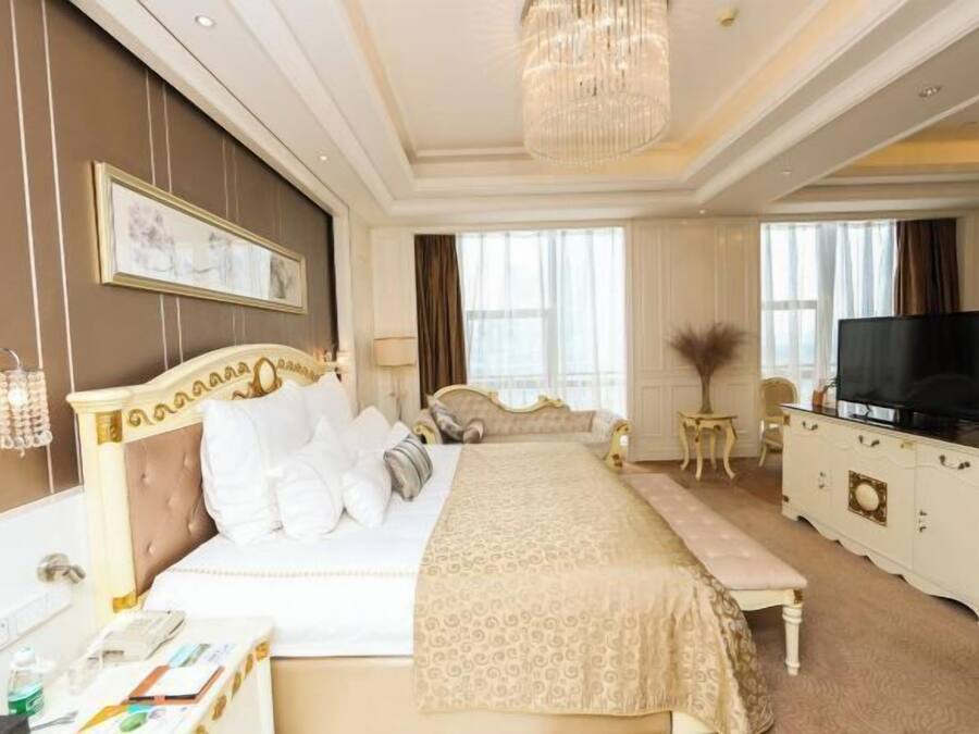 A hotel room with a large bed, a television, a desk with a telephone, and a chandelier.